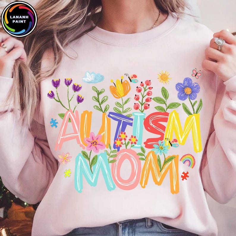 以下が含まれることがあります： 明るいピンク色のスウェットシャツ。カラフルな花柄の文字で「AUTISM MOM」と書かれています。デザインには花、蝶、虹が含まれています。スウェットシャツはクルーネックと長袖です。