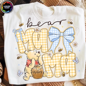 Pode incluir: Camisola branca com gola redonda com um design "bear mama". O design apresenta o Ursinho Pooh, abelhas, borboletas e as palavras "bear mama" em uma fonte l&uacute;dica. As letras "MAMA" est&atilde;o em um padr&atilde;o xadrez amarelo.