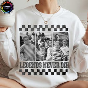 May include: White sweatshirt featuring a black and white image of a group of boys. The image is framed with a checkered pattern and the text "LEGENDS NEVER DIE" is printed below the image. The sweatshirt has a crew neck.