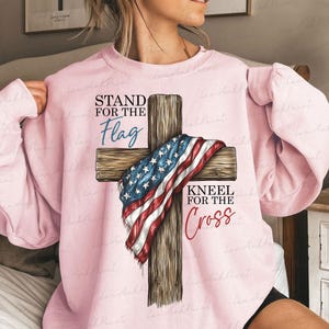 Puede incluir: Sudadera rosa claro con una cruz de madera adornada con una bandera estadounidense. El texto "STAND FOR THE Flag" est&aacute; encima de la cruz y "KNEEL FOR THE Cross" debajo. Una prenda de tem&aacute;tica patri&oacute;tica.