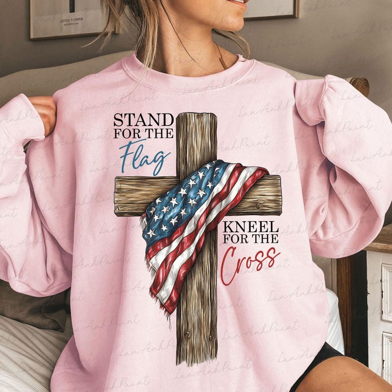 Puede incluir: Sudadera rosa claro con una cruz de madera adornada con una bandera estadounidense. El texto "STAND FOR THE Flag" est&aacute; encima de la cruz y "KNEEL FOR THE Cross" debajo. Una prenda de tem&aacute;tica patri&oacute;tica.
