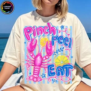 May include: A cream-coloured t-shirt with a vibrant graphic. The design includes a pink lobster, lemon slices, and the words "Pinch Peel Eat" in pink and blue. A small logo is visible in the upper left corner of the shirt.