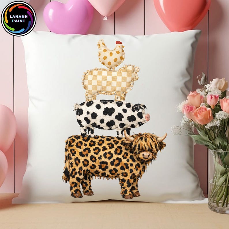 Op de afbeelding: Wit sierkussen met een stapel boerderijdieren: een kip, een schaap, een varken en een koe. De koe heeft een luipaardprint. Het kussen ligt op een houten oppervlak met roze ballonnen en bloemen op de achtergrond.