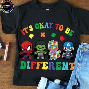 May include: Black t-shirt with the text "It's Okay to Be Different" in rainbow colors. The shirt features cartoon illustrations of Spider-Man, Hulk, a puzzle piece figure, and Captain America. A pair of blue jeans is visible.
