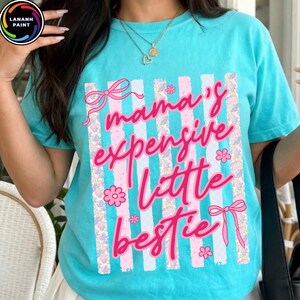 Puede incluir: Camiseta turquesa con la frase rosa "mama's expensive little bestie". El dise&ntilde;o incluye rayas verticales con detalles florales y lazos. La camiseta tiene cuello redondo y mangas cortas.