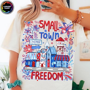 May include: Cream-colored t-shirt with a colorful graphic design. The design features the words "SMALL TOWN FREEDOM" in red and blue, along with illustrations of buildings, flowers, and a water tower. The shirt has a crew neck and short sleeves.