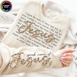 May include: Cream-colored sweatshirt with the word "Jesus" in a leopard print design with yellow bows. The sweatshirt has a printed script with religious text. The brand logo "LANANH PAINT" is in the upper left corner.