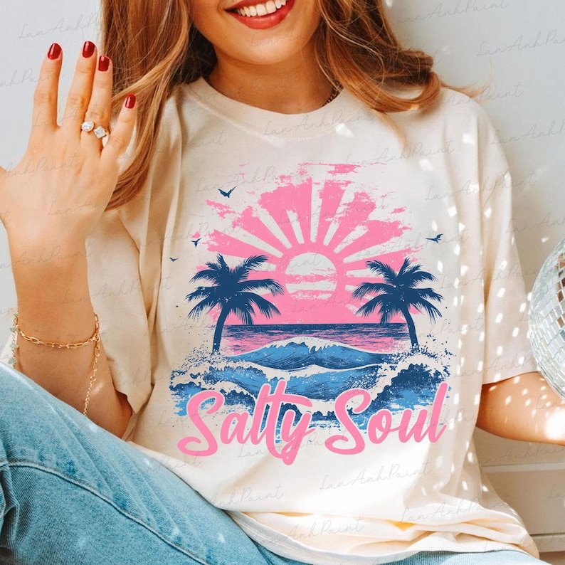 Pu&ograve; includere: Una maglietta bianca con una grafica rosa e blu di un tramonto sull'oceano con palme e il testo "Salty Soul".