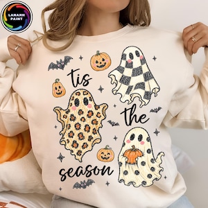 May include: A cream-colored crewneck sweatshirt featuring a graphic print with the text 'tis the season'. The design includes three ghosts with different patterns, including leopard print, black and white checkers, and polka dots, alongside orange pumpkins and small black bats.