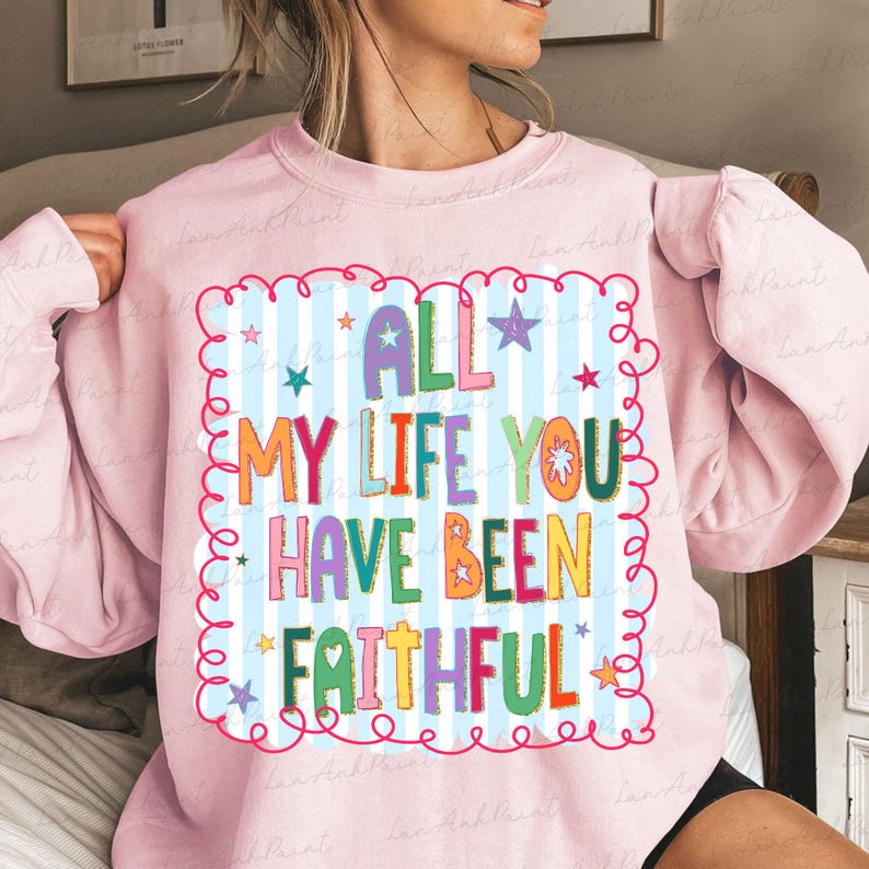 May include: A light pink sweatshirt with the text "ALL MY LIFE YOU HAVE BEEN FAITHFUL" in colorful letters. The text is inside a blue and white striped square with a red curly border and star accents. The sweatshirt has long sleeves.
