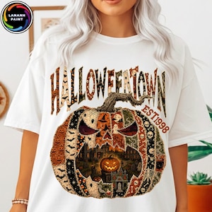 May include: A white t-shirt featuring a large graphic of a pumpkin composed of various Halloween-themed patterns. The text HALLOWEENTOWN EST 1998 is printed above the pumpkin, which contains a detailed scene of a haunted house and a smaller pumpkin.