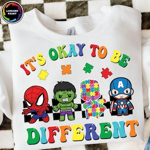 May include: White sweatshirt with the text "IT'S OKAY TO BE DIFFERENT" in rainbow colors. Features cartoon illustrations of Spider-Man, Hulk, a puzzle piece figure, and Captain America. The sweatshirt has a crew neck.
