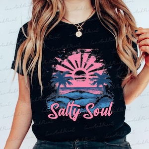 Pu&ograve; includere: Una maglietta nera con una grafica di tramonto rosa e blu e il testo "Salty Soul" in rosa.