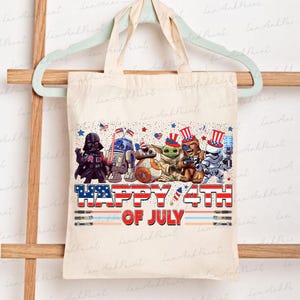 K&ouml;nnte beinhalten: Beige Tragetasche mit einem farbenfrohen Grafikdesign. Es zeigt Star Wars-Charaktere wie Darth Vader, R2-D2 und Grogu, sowie den Text "HAPPY 4TH OF JULY". Die Tasche ist aus Stoff gefertigt.
