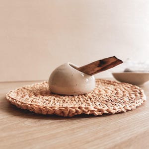 以下が含まれることがあります： 滑らかなオフホワイトのセラミック製ホルダー。丸みを帯びた形状で、斜めに開いた穴が一つあります。ホルダーの穴には茶色い質感のある木片が差し込まれており、丸く編まれた明るい茶色のストローマットの上に置かれています。