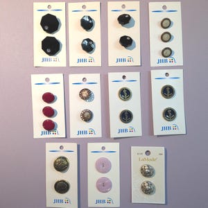 Könnte beinhalten: Eine Sammlung von elf Knopfsets der Marken JHB und LaMode auf weißen Präsentationskarten. Die Knöpfe variieren in Form, Farbe und Design, darunter schwarze achteckige, kastanienbraune, metallische und silberfarbene runde Modelle.