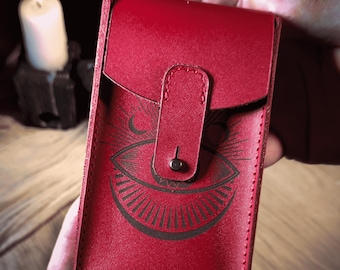 May include: A red leather card case with a flap closure and a button clasp. The case features a black graphic of an eye with a crescent moon above it. The case is held in a person's hands.