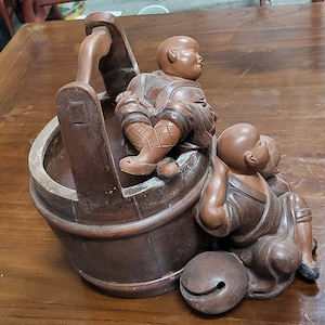 May include: A wooden sculpture of two figures sitting on a wooden bucket. The figures are carved in a traditional style and have a brown finish.