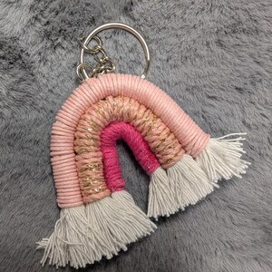 May include: A pink, white, and gold rainbow keychain with white tassels. The keychain is made of yarn and has a silver metal ring.