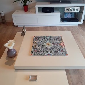 Peut inclure: Salon moderne avec une table basse blanche ornée d'une mosaïque en son centre. Un petit cendrier carré est posé sur la table inférieure. Un meuble TV blanc avec une télévision et une plante en pot rouge sont en arrière-plan.