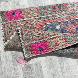 May include: A colorful, rectangular rug with a distressed, vintage appearance. The rug features a pattern of pink, blue, and green geometric shapes on a neutral background. The edges are fringed.