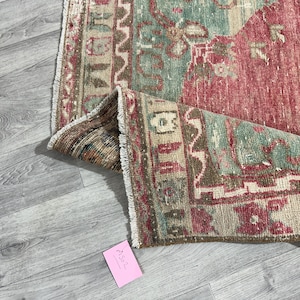 Puede incluir: Una sección de una alfombra vintage con un aspecto desgastado y descolorido. La alfombra presenta una combinación de tonos rojos, verdes y beige. Los bordes están deshilachados y la alfombra está parcialmente enrollada, revelando sus intrincados patrones y texturas.