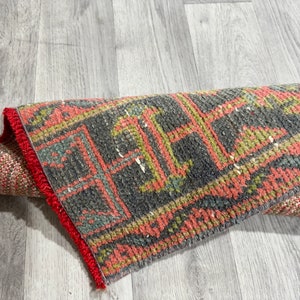 Pode incluir: Um tapete vermelho e cinza enrolado com uma borda de franjas. O tapete tem um design geométrico com cores vermelho, cinza e verde.