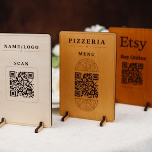 Może przedstawiać: Trzy drewniane tabliczki z kodami QR i tekstem. Pierwsza tabliczka zawiera napis "NAME/LOGO" i "SCAN". Druga tabliczka zawiera napis "PIZZERIA MENU". Trzecia tabliczka zawiera napis "Etsy Kup online".