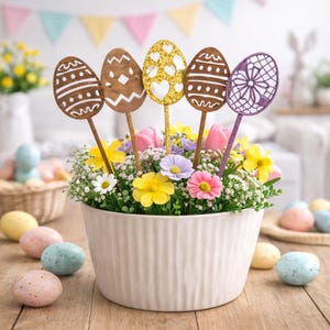 May include: A white ceramic pot filled with colorful flowers and decorative Easter egg picks. The egg picks are made of wood and glitter, with various designs. Several pastel-colored Easter eggs are scattered around the pot on a wooden surface.