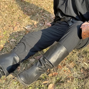 Puede incluir: Un par de botas de cuero negro con un dobladillo. Las botas las lleva puestas una persona sentada en una superficie cubierta de hierba. La persona lleva pantalones negros y una camisa negra con un cintur&oacute;n de cuero marr&oacute;n.