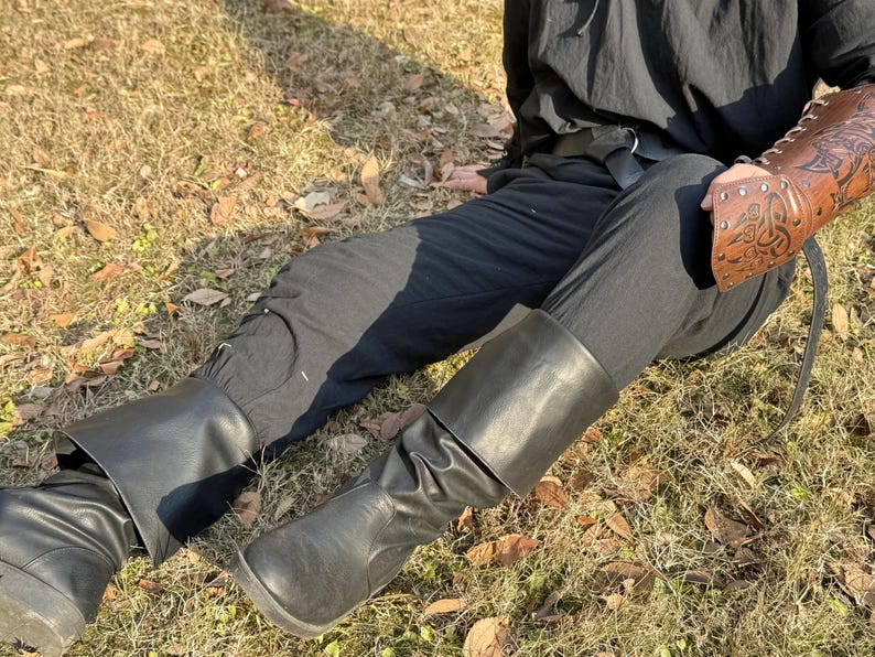 Puede incluir: Un par de botas de cuero negro con un dobladillo. Las botas las lleva puestas una persona sentada en una superficie cubierta de hierba. La persona lleva pantalones negros y una camisa negra con un cintur&oacute;n de cuero marr&oacute;n.