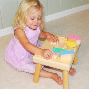 May include: A young child sits on a wooden stool with colorful letters spelling out their name. The stool is made of natural wood and has four legs. The letters are painted in pastel colors and are arranged in a row on the top of the stool.