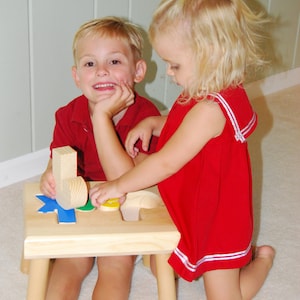 May include: Two children, a boy and a girl, are playing with a wooden puzzle toy. The boy is sitting on a small wooden stool and the girl is kneeling on the floor. The puzzle has different colored shapes that fit into the corresponding holes. The boy is wearing a red shirt and the girl is wearing a red dress with white stripes.