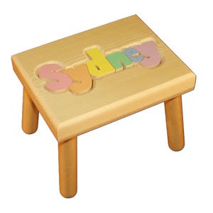 May include: A wooden step stool with the name "Sydney" spelled out in colorful letters. The stool is made of natural wood and has a light brown finish.