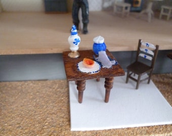 A one 1/4 inch scale tiny table with a hand painted chair.There is an oil lamp,a blue and white jug and plate with a jam tart and a napkin.