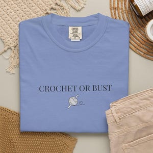 May include: A periwinkle blue crew neck t-shirt featuring the black text CROCHET OR BUST centered on the chest. Below the text is a small black line drawing of a ball of yarn with a crochet hook and a tiny heart outline attached to the yarn.