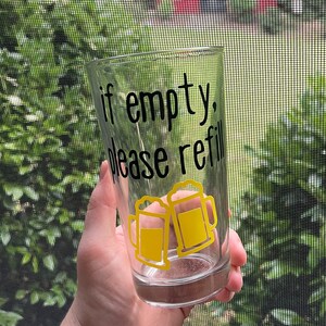 Puede incluir: Un vaso de vidrio transparente con el texto en negro "if empty, please refill" y dos iconos gráficos amarillos de jarras de cerveza. El diseño está impreso en la parte frontal del recipiente cilíndrico, que tiene una base plana y un borde liso.