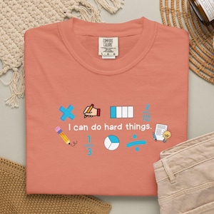 May include: A coral-colored t-shirt featuring the text "I can do hard things." surrounded by various math-themed graphics including a multiplication sign, a hand holding a pencil, fractions, a division sign, a pencil, and a lightbulb.
