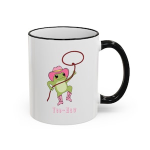 May include: A white ceramic mug with a black rim and handle, featuring a graphic of a green frog wearing a pink cowboy hat and pink boots, holding a lasso. Below the frog, the text YEE-HAW is printed in pink letters on the white surface.