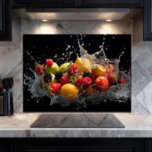 Op de afbeelding: Een keukenachterwand met een dynamisch beeld van fruit en water. De afbeelding toont een kom met fruit, waaronder aardbeien, frambozen, sinaasappels en appels, met water dat eromheen spettert. De achtergrond is zwart, wat een opvallend contrast creëert.