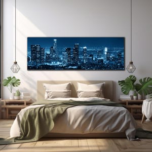 May include: A large rectangular canvas print of a cityscape at night, dominated by tall buildings illuminated in shades of blue. The print hangs above a bed with beige bedding and a green throw. The room is decorated with plants and pendant lights.