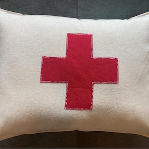 May include: A white pillow with a red cross sewn on the front. The cross is outlined in white stitching.