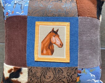 May include: A square patchwork throw pillow featuring a central panel with a brown horse head illustration framed in yellow. The surrounding fabric blocks include brown corduroy, blue denim, brown paisley print, faux cowhide, and blue sky print.