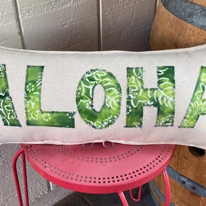 May include: A white pillow with the word "ALOHA" spelled out in green fabric with a floral pattern. The pillow is sitting on a red stool.