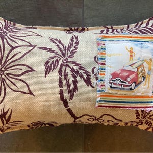 May include: A beige pillow with a brown palm tree print and a colorful fabric patch featuring a vintage car and surfers. The patch has a white background with colorful stripes around the edges.