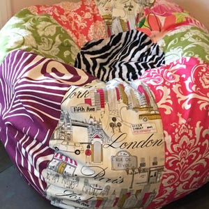 May include: A large, round beanbag chair with a patchwork design. The fabric features various patterns, including floral, zebra print, and cityscapes with text that reads "London", "Paris", and "Fifth Ave". The color palette includes pink, green, purple, and white.