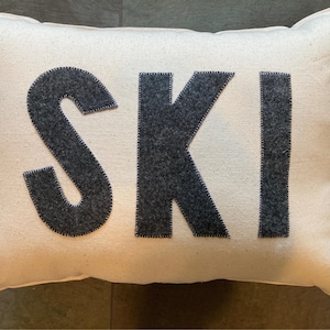 May include: Rectangular off-white pillow with the word "SKI" in dark gray felt letters. The letters are stitched onto the pillow. The pillow has a soft, textured appearance, suggesting a cozy home decor item.
