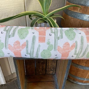 May include: A white lumbar pillow with a green and pink cactus print. The pillow is sitting on a wooden crate.