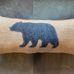 May include: A rectangular lumbar pillow featuring a textured tan burlap fabric cover. Centered on the front is a dark gray felt applique in the shape of a walking bear, showcasing a simple silhouette and a soft, matte finish on the fabric surface.