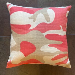 May include: A square throw pillow featuring a camouflage pattern in shades of coral pink and light beige. The fabric has a visible woven texture throughout the surface, creating a consistent look across the entire front of the pillow.